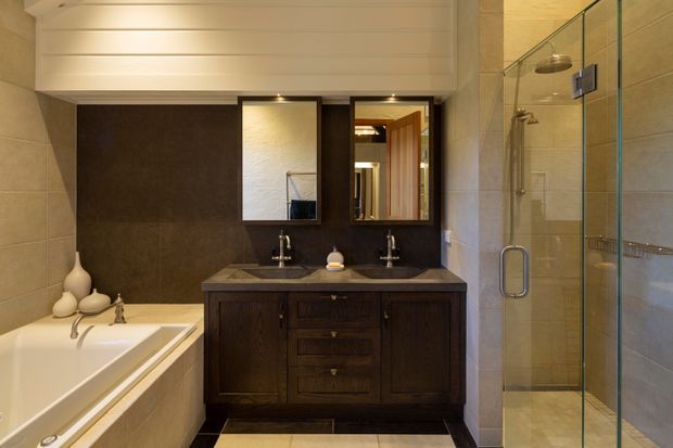 A warm, traditionally styled bathroom with a double vanity in dark timber cabinetry, two framed mirrors, a white bathtub, and a glass-enclosed walk-in shower. Beige tiled walls and a white horizontal-panel ceiling create a refined, earthy palette.