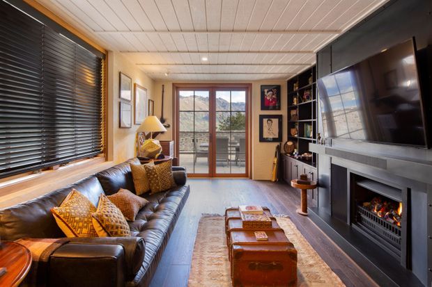 A compact, warmly furnished living room with a dark leather sofa, patterned cushions, a central leather trunk coffee table, built-in shelving, a large wall-mounted television, and a contemporary fireplace surround. Timber-framed glazing and a glass door open to mountain views, while a white-painted timber-lined ceiling, dark timber floor, and warm neutral walls create a cozy lodge-like atmosphere.