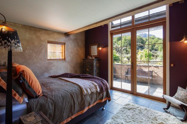 A warm, rustic bedroom with a large bed dressed in layered grey and burnt-orange textiles, a textured plaster wall, dark timber beams, and tall timber-framed glazed doors opening to a terrace. Natural light, mountain greenery, and a small outdoor seating area are visible beyond the glazing. The room also includes a vintage-style dresser, pendant lighting, and a plush light rug.