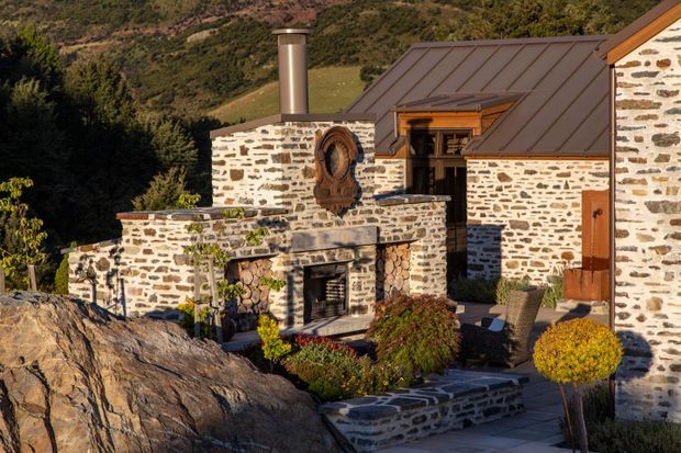 Exterior view of a rustic stone house set into a mountainous, wooded landscape. The building features pale irregular stone masonry walls with dark mortar, a dark standing-seam pitched roof, timber-framed openings and trim, a prominent stone chimney, and an outdoor stone fireplace or hearth. Stone-paved steps and terraces with clipped shrubs lead through the foreground.