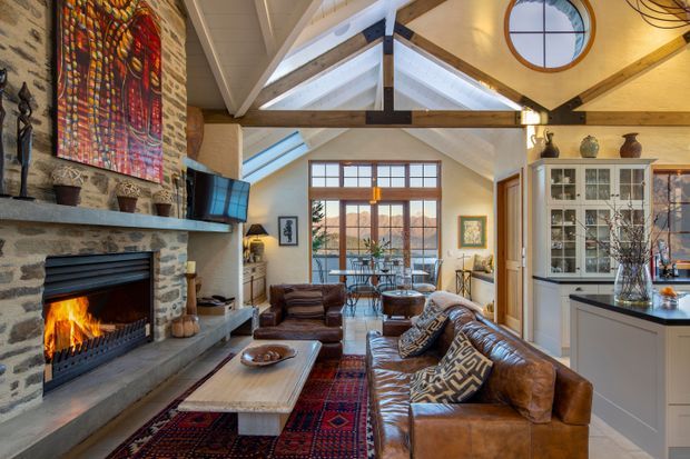 Warm, rustic-contemporary open-plan living room with exposed timber roof framing, high vaulted ceiling, a large stone fireplace, leather sectional seating, patterned rugs and cushions, and a dining area beyond. A glazed built-in display cabinet and kitchen joinery are visible to the right, with abundant natural light from tall windows and clerestory glazing.