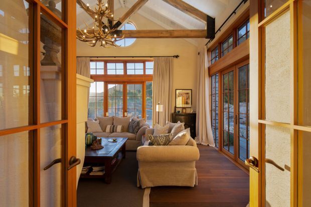 A warm, rustic living room viewed through glazed timber double doors. The room features exposed timber beams and rafters, extensive timber-framed glazing, dark timber flooring, upholstered sofas and armchairs arranged around a coffee table, and a sculptural antler-style chandelier. Large windows and doors provide abundant natural light, while textured neutral walls and curtains create a cosy lodge-like atmosphere.