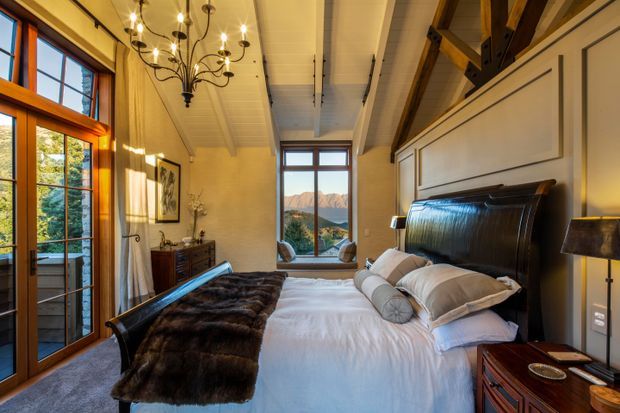 A warm, elegant bedroom with a large bed positioned beneath exposed timber roof framing. The room features white wall panelling, a vaulted white-lined ceiling, rich timber-framed windows and glazed doors, dark wood furniture, and an ornate multi-arm chandelier. Mountain scenery is visible through the glazing.