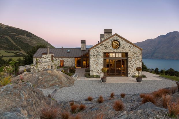 A stone-clad mountain residence positioned above a deep blue lake, with a prominent gabled central pavilion, multiple chimneys, large timber-framed glazed doors, and a circular feature window. The landscaped foreground uses gravel, exposed rock, and sparse ornamental grasses, while rugged mountains surround the property.
