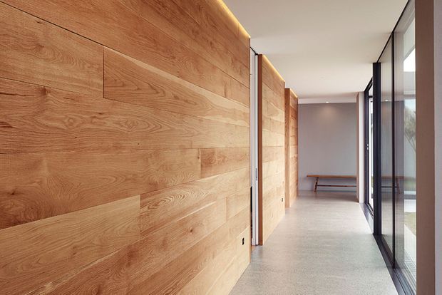 A long, minimalist interior hallway with a full-height warm timber feature wall made from wide horizontal boards, concealed linear lighting at the ceiling edge, a smooth light terrazzo or concrete floor, and extensive dark-framed floor-to-ceiling glazing along the right side. A simple timber bench is visible at the far end, creating a calm, refined circulation space with strong natural light.