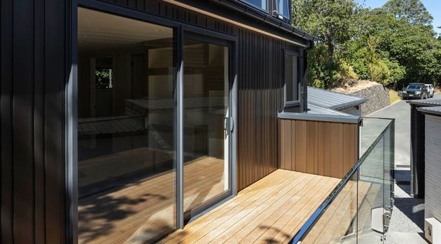 Exterior view of a contemporary multi-level home with dark vertical cladding, large glazed sliding doors, and a timber deck enclosed by a frameless glass balustrade. The deck overlooks a sloped driveway and neighbouring roof forms, with dense trees beyond. Interior living space is visible through the glazing.