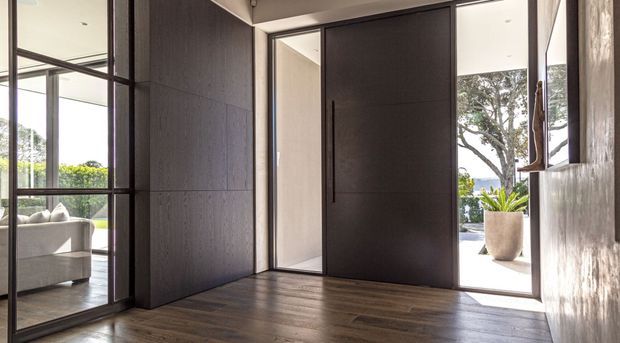 A refined modern entryway with dark timber flooring, a large pivot-style charcoal door, and expansive black-framed glazing. The open doorway reveals a bright outdoor terrace with a potted plant, while the glazing reflects or provides views toward an adjacent living area with a light sofa. Textured dark wall panelling and minimal detailing create a sleek, high-contrast entrance.