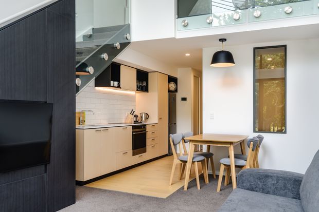 Contemporary open-plan kitchen and dining area beneath a mezzanine or loft. The compact kitchen features pale timber cabinetry, dark benchtops and appliances, under-cabinet lighting, and black-framed details. A small timber dining table with upholstered chairs sits beside a tall glazed window, while a dark feature wall and staircase with glass balustrade define the adjacent living area. The space is bright, compact, and efficiently planned with a modern urban feel.