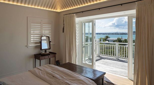 A bright coastal bedroom opening onto a timber balcony or terrace, with a partial water view beyond. The room features a bed, dark wood console table with mirror, plantation shutters, full-height glazed doors, cream curtains, and a distinctive ribbed ceiling with concealed warm lighting.