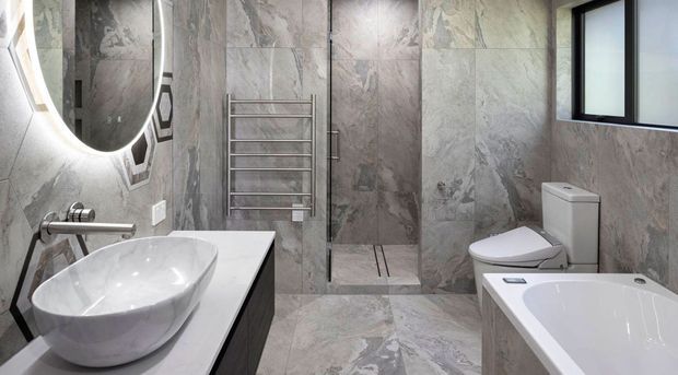 Contemporary bathroom featuring full-height grey marble-look wall and floor tiles, a freestanding vessel basin on a dark vanity, illuminated round mirror, chrome tapware, heated towel rail, frameless glass shower, toilet and a white bathtub. A black-framed window provides natural light.