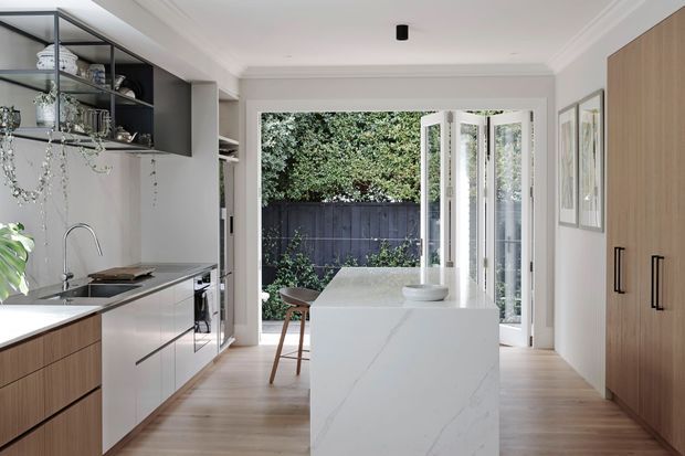 Bright contemporary kitchen with a white waterfall island, pale timber flooring, light cabinetry, black-framed open shelving, and full-height bi-fold glazing opening to a leafy outdoor area. A tall timber pantry or storage wall lines the right side, while pendant lighting and framed artwork add restrained decorative detail.