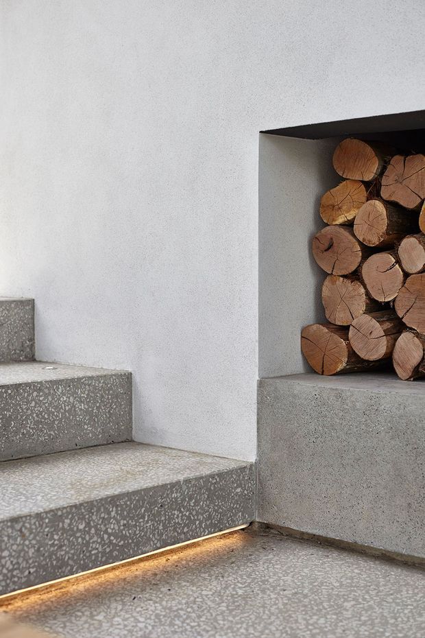 Close-up architectural detail of a minimalist concrete stairway with terrazzo-like aggregate finishes and warm recessed lighting beneath the lower step. A built-in wall niche beside the stairs is filled with stacked firewood logs, creating a natural contrast with the cool grey surfaces.