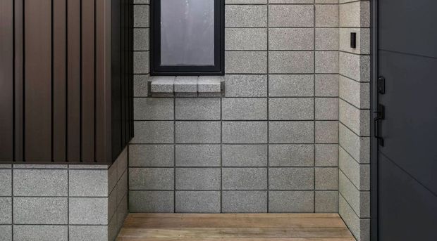 A compact modern entry porch featuring light grey concrete block walls, a dark vertical timber-clad facade section, a dark contemporary entry door, a narrow window, and warm timber decking.