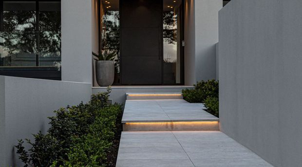 Modern residential entrance with a wide tiled walkway and two raised steps illuminated by warm recessed strip lighting. Smooth white rendered walls, dark framed glazing, a large dark entry door, and neatly clipped shrubs create a restrained contemporary facade.