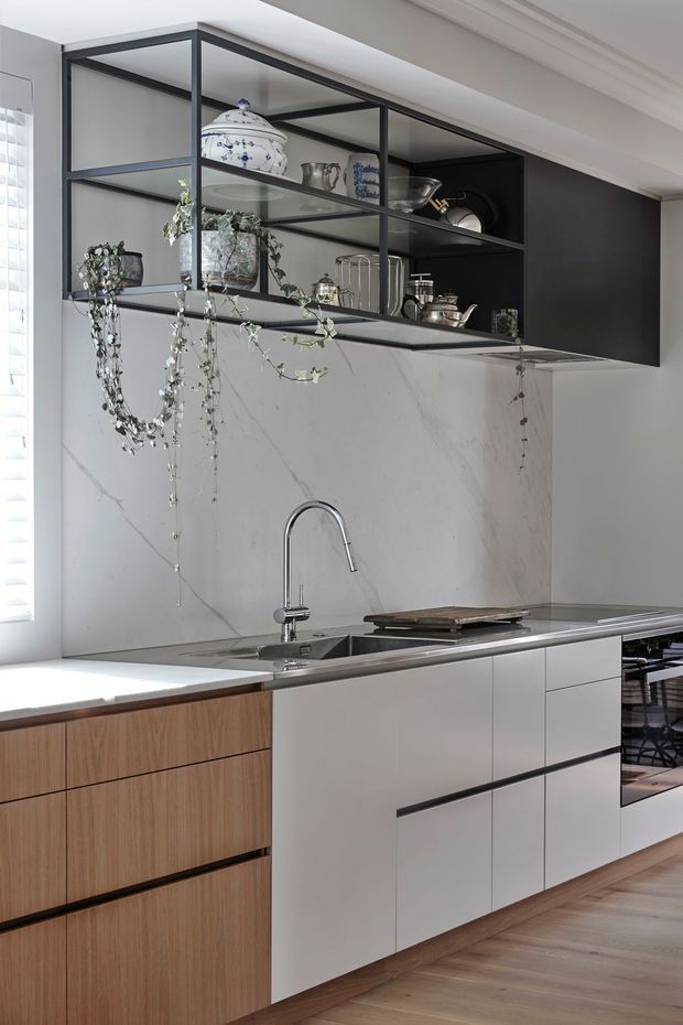 A refined contemporary kitchen featuring white and timber cabinetry, a large marble-look splashback, stainless steel sink and gooseneck tap, and a dark stone or metal benchtop. Black-framed glass display shelving spans the upper wall, showcasing ceramics, glassware and trailing plants. The kitchen opens toward a dining area visible at right, with warm timber flooring and abundant natural light from the window at left.