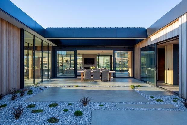 Contemporary single-storey courtyard-style residence viewed from an outdoor paved forecourt. The composition features crisp dark metal roof planes, extensive floor-to-ceiling glazing, vertical timber cladding, and a central glazed living and dining area opening onto minimalist gravel landscaping with concrete paths. A dining table and chairs are visible inside, with a fireplace or media feature on the rear wall.