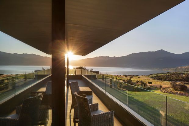 A covered outdoor terrace or balcony overlooking a lake, mountain range, and broad valley. The space features a projecting timber-toned roof, transparent glass balustrades, dark structural posts, and woven outdoor dining chairs around a table. Warm low sunlight creates a serene, scenic atmosphere.