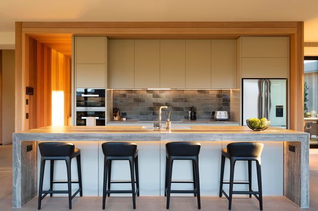 A warm, contemporary kitchen viewed straight on, centred around a large island with four black bar stools. The kitchen features light beige flat-panel cabinetry, a grey-brown brick-look tiled splashback, a pale stone island benchtop, integrated appliances, timber-lined wall and ceiling details, and a large stainless-steel refrigerator. Timber-framed openings connect the kitchen to adjacent spaces.