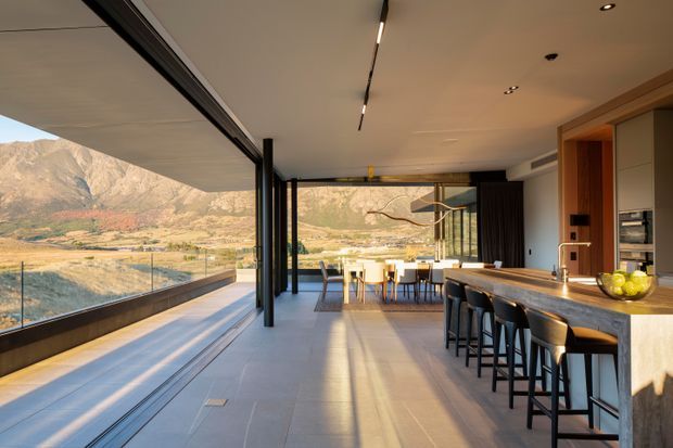 A spacious, contemporary open-plan kitchen and dining area with floor-to-ceiling retractable glazing opening onto a dramatic mountainous landscape. The kitchen features a long stone island with integrated sink and timber cabinetry, while a dining table sits beyond beneath a sculptural pendant light. Dark-framed glazing, linear ceiling lighting, and polished tiled flooring create a refined indoor-outdoor connection.