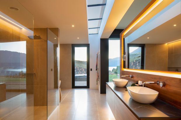 Contemporary double-basin bathroom with a large illuminated mirror, floating dark timber vanity, vessel basins, frameless glass shower, and full-height glazing overlooking a mountainous landscape. A linear skylight and recessed ceiling lighting add brightness and a strong sense of openness.