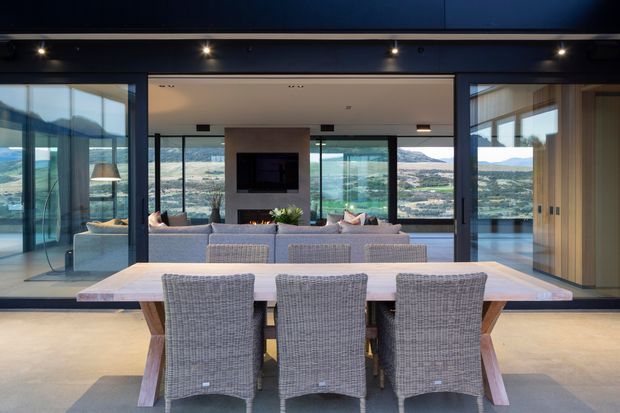 A contemporary open-plan dining and living area with a long light-toned dining table and three woven dining chairs in the foreground. Dark framed floor-to-ceiling glazing and sliding doors open the room to expansive mountain and valley views. A lounge with a television, fireplace, and low furnishings is visible beyond, with warm timber cabinetry and a dark ceiling creating contrast.