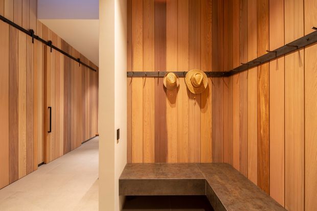 Warm, narrow dressing and entry corridor lined with vertical timber cabinetry and wall panelling. A black rail runs along the walls with hats hanging above an L-shaped dark timber or stone bench, while a pale floor and cream structural partition brighten the space.