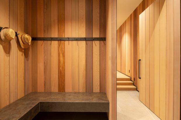 Warm contemporary timber-lined entry or hallway with extensive vertical wood wall panelling, an integrated dark bench seat, black rail with woven hats, and a bright adjoining corridor with timber stairs and concealed cabinetry. The space has a refined, natural and softly illuminated atmosphere.