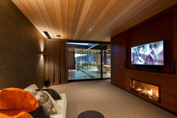 A warm, dark-toned media room with a large timber-lined feature wall incorporating a television, gas fireplace and low storage cabinetry. A sectional sofa sits in the foreground, while full-height glazing and curtains open toward a bright adjacent outdoor or living area. The ceiling is clad in linear timber boards, and integrated wall lighting creates a cozy atmosphere.