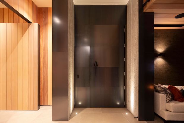 A warm, refined interior entry zone featuring a tall dark timber door set within a textured stone or concrete surround. Vertical timber wall panelling and integrated lighting frame the doorway, while a partially visible lounge with a sofa, dark textured wall, timber ceiling, and recessed lighting appears to the right.