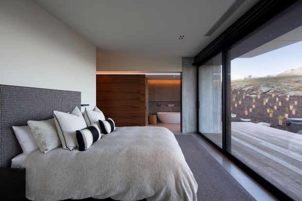 A contemporary bedroom with a large upholstered bed, layered neutral bedding and monochrome accent cushions. Full-height black-framed glazing opens the room visually to a timber deck and landscaped rocky terrain. A warm timber feature wall and recessed indirect lighting lead toward a partially visible ensuite bathroom with a freestanding white bathtub.