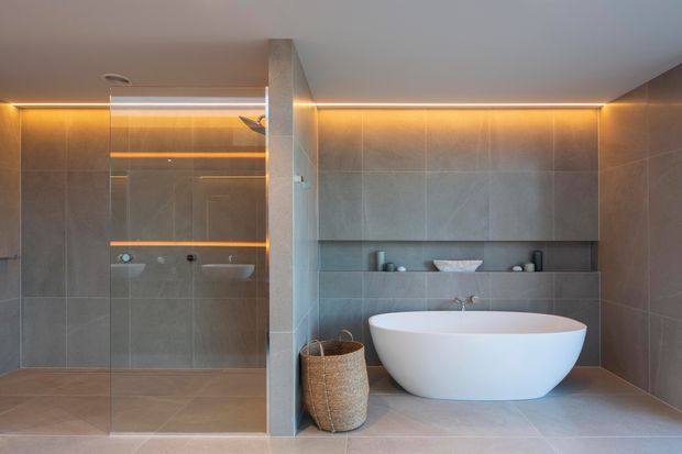 Contemporary bathroom featuring a freestanding oval bathtub, walk-in shower with frameless glass, large-format grey stone-look wall and floor tiles, recessed shelving, and warm concealed LED lighting.