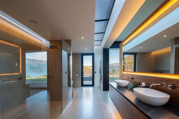 Contemporary luxury bathroom with a long double vanity, two vessel basins, dark timber cabinetry, illuminated mirror, freestanding-style bath partially visible at left, and large-format pale floor and wall tiles. Floor-to-ceiling glazing and a central glass roof panel bring in natural light, with a dramatic mountain and lake landscape visible beyond.