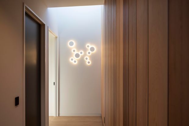 A narrow, warmly lit residential hallway with light timber flooring, a full-height vertical timber-panelled wall or built-in joinery on the right, and dark timber door frames on the left. A sculptural cluster of circular wall lights forms a feature on the pale end wall, while a bright opening or skylight adds gentle illumination.