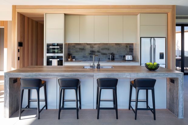 Contemporary open-plan kitchen with a large stone island and four dark bar stools. The kitchen features light cream flat-panel cabinetry, warm timber wall and ceiling detailing, a grey textured tile splashback, integrated appliances, and a stainless-steel refrigerator. Glazing and a dining or living area are visible beyond the kitchen.