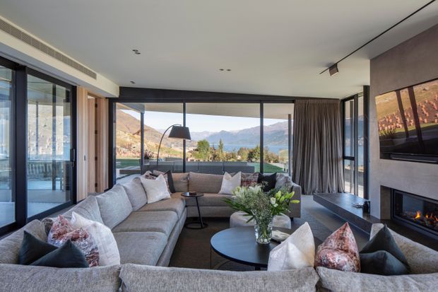 A refined contemporary living room with a large grey sectional sofa arranged around layered round coffee tables, facing a wall-mounted television and fireplace. Floor-to-ceiling glazing opens to a mountain landscape, with full-height curtains, timber wall detailing, and a neutral, textured interior palette.