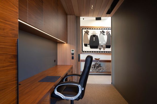 Compact, highly tailored home office with full-height warm timber wall panelling and integrated cabinetry, a built-in timber desk, dark inset shelving, linear LED lighting, and a black ergonomic office chair. A patterned artwork and shelving are visible at the far end, while dark doors or wall surfaces frame the space. The room has a refined, warm modern character with a narrow, corridor-like layout.