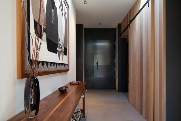 A refined contemporary hallway or entry passage with a long dark timber console, sculptural vase, and large abstract artwork on the left. The right side features full-height vertical timber wall panelling and concealed cabinetry or doors, while a dark glazed door anchors the far end. Recessed ceiling lights and large-format neutral floor tiles create a warm, minimalist transition space.