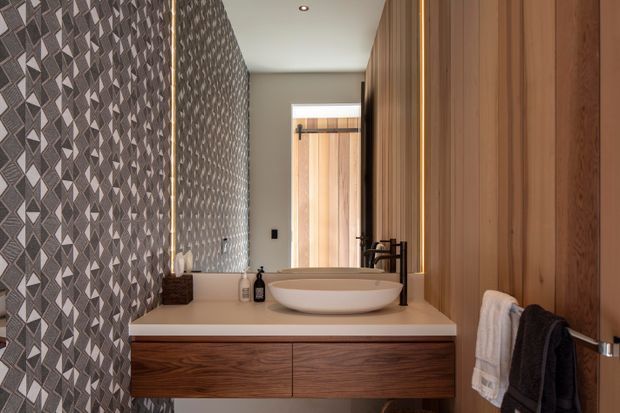 A compact, refined bathroom viewed toward a floating timber vanity with a white stone benchtop, round vessel basin, and matte black tapware. The left wall features a bold geometric patterned tile, while the right side has warm vertical timber wall panelling and a timber door. A mirror reflects the patterned wall and vanity area, and dark and white towels hang beside the basin.