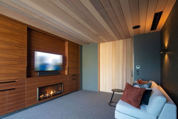 A cozy contemporary media room with a wall-mounted television, built-in dark timber cabinetry, and a linear electric fireplace. The room features a full-height timber-panelled ceiling, vertical light timber wall cladding, charcoal textured wall finishes, recessed lighting, and a pale sectional sofa with warm-toned cushions.
