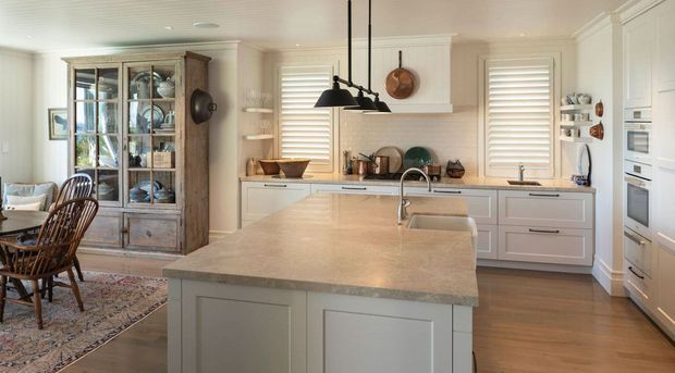 A bright, traditional-style kitchen with a large central island, white shaker cabinetry, pale stone benchtops, timber flooring, plantation shutters, pendant lighting, and a freestanding glass-front timber display cabinet. A dining area is visible at the left edge of the image.