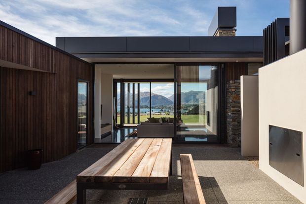 A contemporary outdoor entertaining courtyard framed by dark vertical timber-clad walls and a low flat-roofed residence. A substantial timber dining table and bench occupy the foreground, while full-height glazed doors open toward an interior living area and a dramatic mountain-and-water landscape beyond. A built-in white rendered outdoor fireplace is visible on the right, with stone cladding around an adjacent vertical element.
