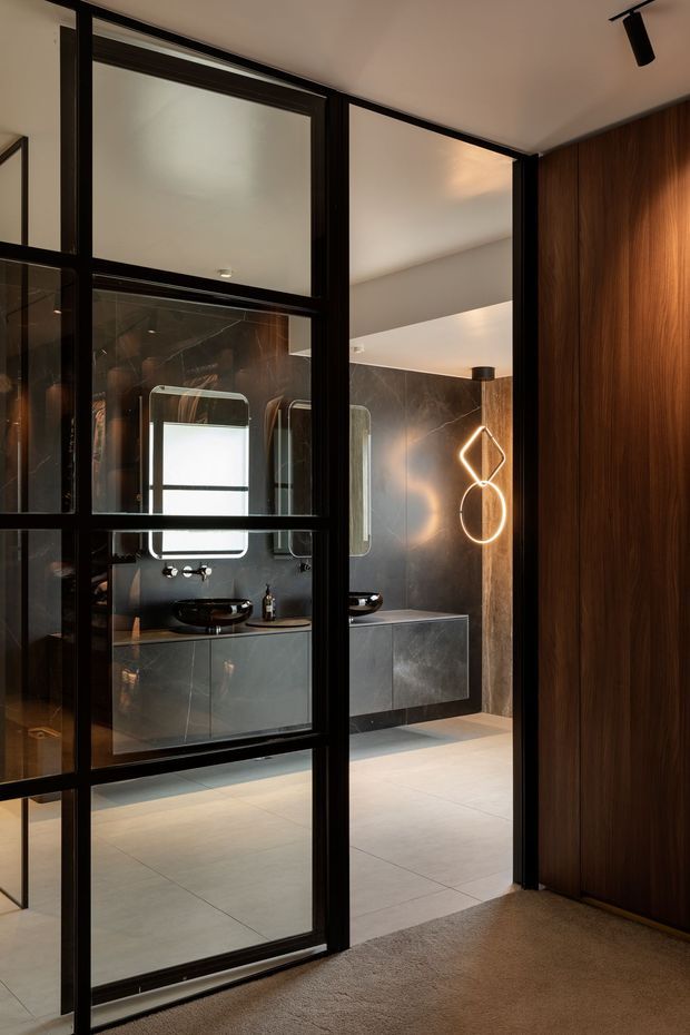 A sophisticated modern bathroom viewed through black-framed glazed steel-style partitions and doors. The room features dark marble-look wall surfaces, a floating grey vanity with black vessel basins, illuminated mirrors, warm timber wall panelling, pale large-format floor tiles, and sculptural wall lighting. The foreground includes carpeted flooring and a dark timber wall or door.