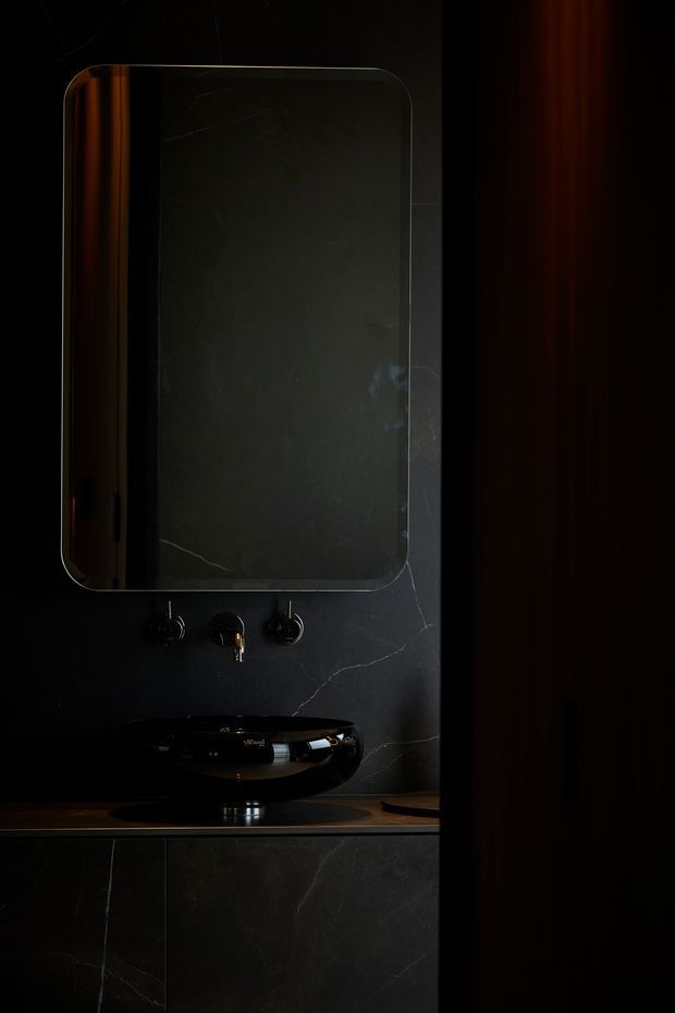 A moody, dark bathroom vanity composition featuring a rounded-rectangle mirror, black wall-mounted basin with exposed tapware, and dark marble-look wall and cabinetry surfaces. Warm timber tones appear at the vanity counter and along the right side, with dramatic low-key lighting.