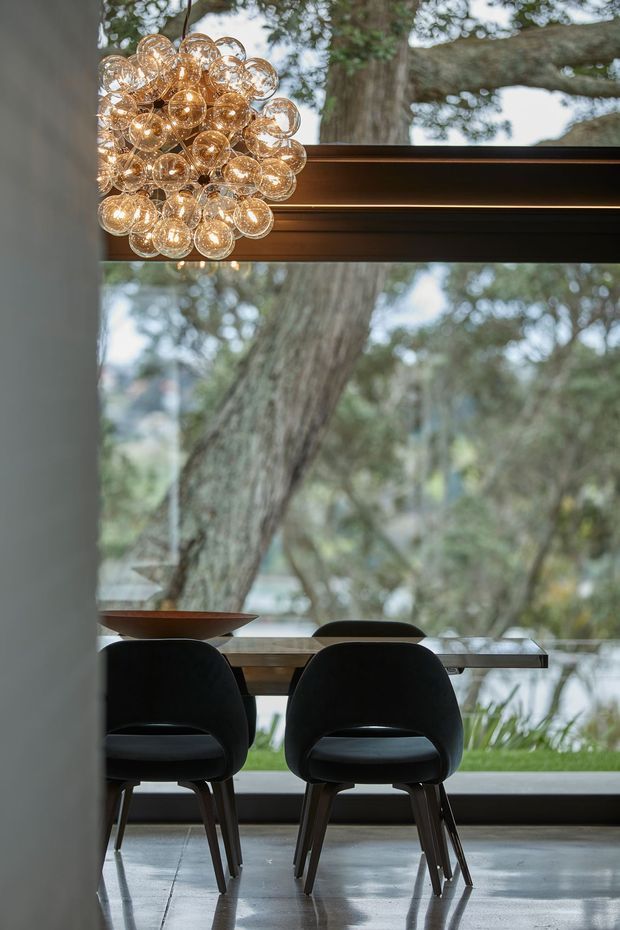 A refined dining area beside expansive floor-to-ceiling glazing, with a dark wood-framed window, polished concrete floor, slender dining table, sculptural black chairs, and a dramatic cluster pendant composed of many clear glass globes. Mature trees are visible outside.