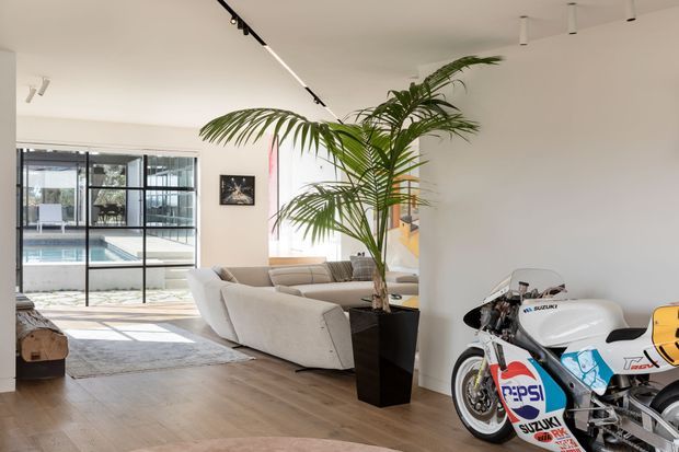 Bright contemporary open-plan living room with pale timber flooring, light upholstered sofas, white walls and ceiling, track lighting, and a large indoor palm. Full-height black-framed glazing opens views toward a swimming pool and outdoor area. A prominently displayed Suzuki racing motorcycle adds a distinctive accent to the otherwise calm, minimalist interior.