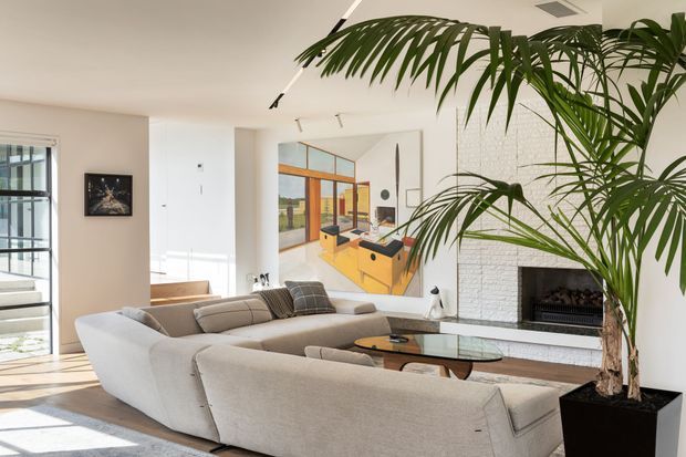 Bright contemporary living room with a large curved light-upholstered sectional sofa, glass coffee table, white textured brick fireplace, timber flooring, and expansive glazing. A large indoor palm adds greenery, while a colourful abstract artwork provides a focal accent. The room has clean lines, restrained detailing, and abundant natural light.