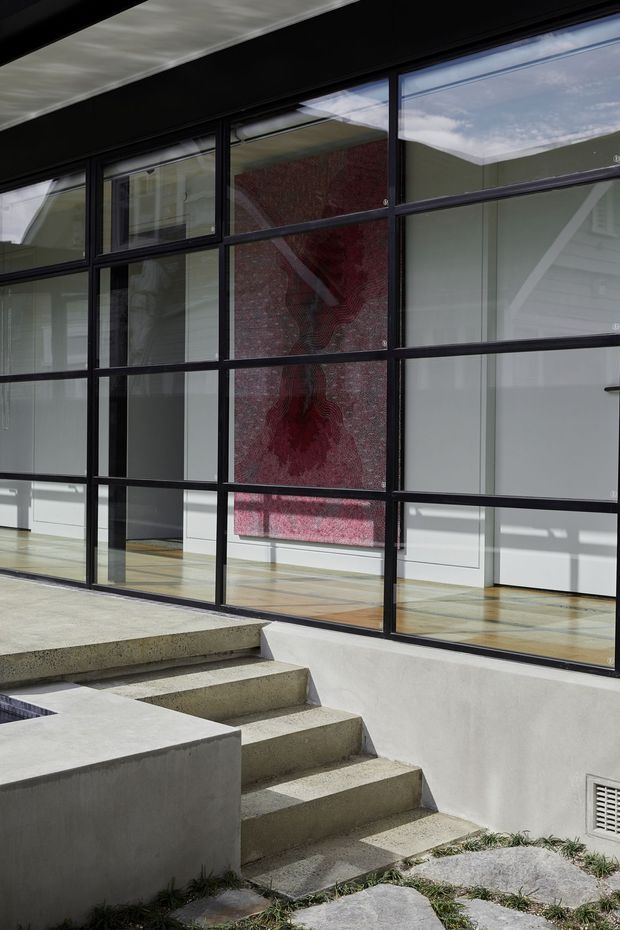 A modern architectural entry or courtyard stairway viewed from outside, with broad raw concrete steps leading to a glazed façade. Large black-framed grid windows reveal a bright interior with timber flooring and a striking red wall artwork or feature surface. The composition emphasizes strong geometric lines, concrete construction, extensive glazing, and indoor-outdoor connection.