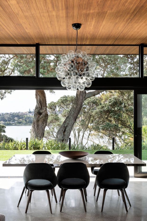 A refined dining area with a long glossy white dining table, black upholstered dining chairs, and a sculptural cluster-glass pendant light. Full-height black-framed glazing opens the room to views of mature trees, landscaped gardens, and a body of water. The ceiling is clad in warm timber, contrasting with the polished concrete floor and dark window framing.