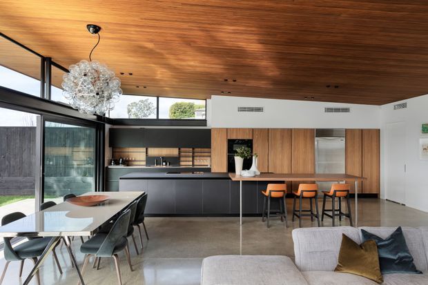 Open-plan contemporary kitchen, dining and living area with a polished concrete floor, extensive timber cabinetry, a dark kitchen island, and a dramatic timber-lined ceiling. The space features floor-to-ceiling glazing, a sculptural pendant light, a dining table with dark chairs, and a compact breakfast bar with orange stools.