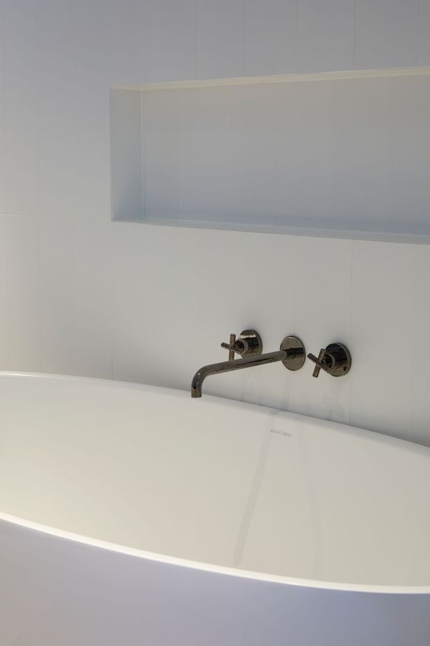 Minimalist bathroom detail featuring a freestanding white bathtub with wall-mounted chrome tapware and a recessed open shelf above. The walls and tub are predominantly white, creating a clean, understated composition.
